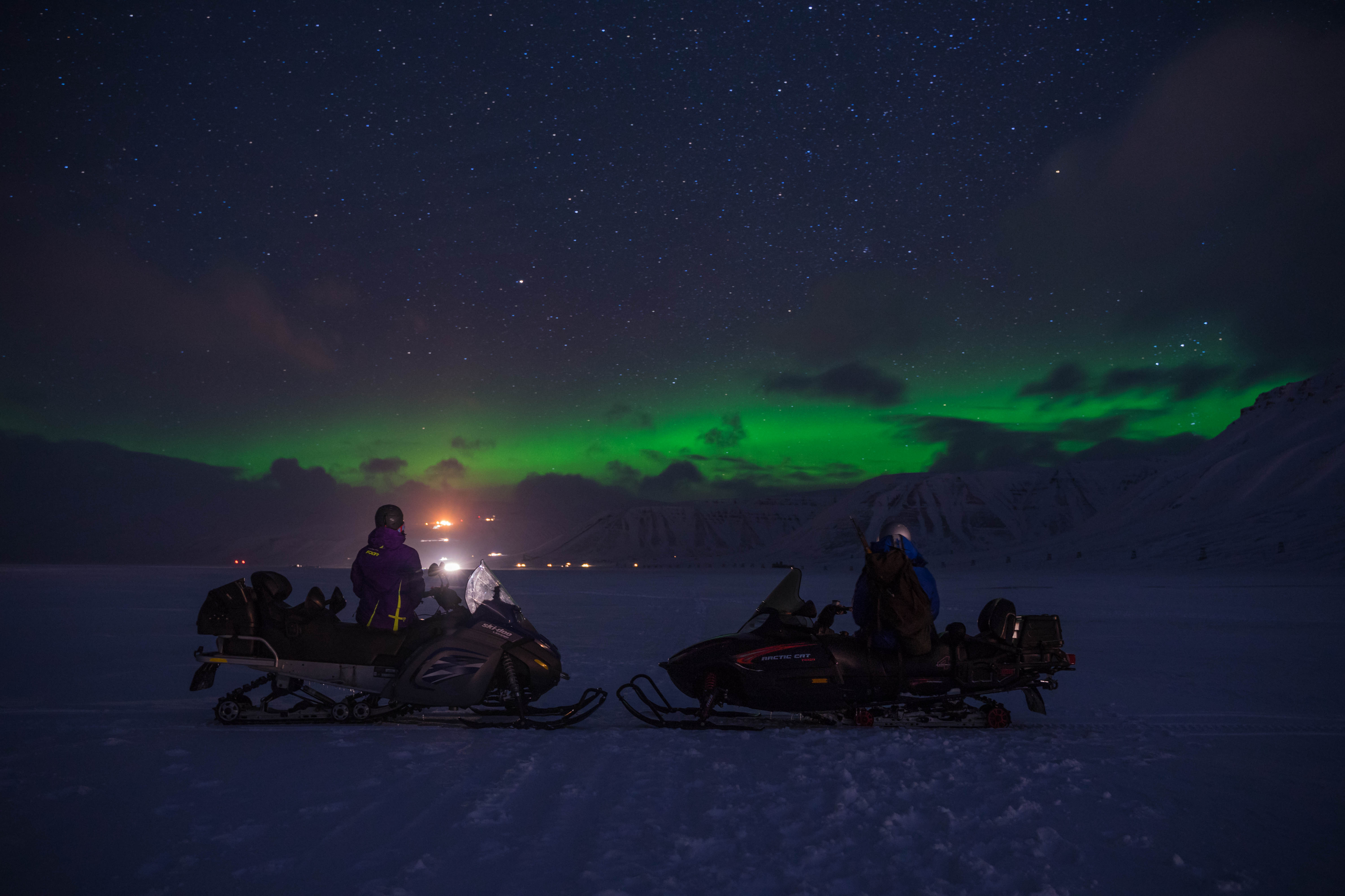 To persons on snowmobiles looking out across a landscape with northern lights in the skies in the background